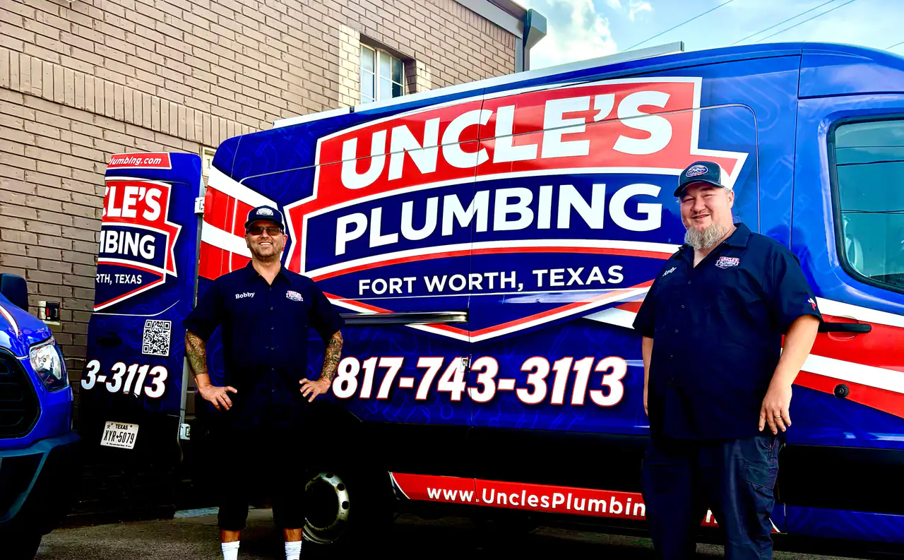 Two Uncle's Plumbing technicians in navy uniform shirts standing on either side of a branded blue service van outside the shop
